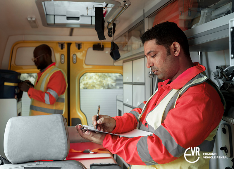 Two emergency workers in safety jackets inside a service vehicle, one writing on a clipboard, one organizing gear.