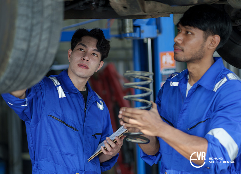Two mechanics in blue uniforms inspecting a vehicle and handling a coil spring in an auto repair shop.